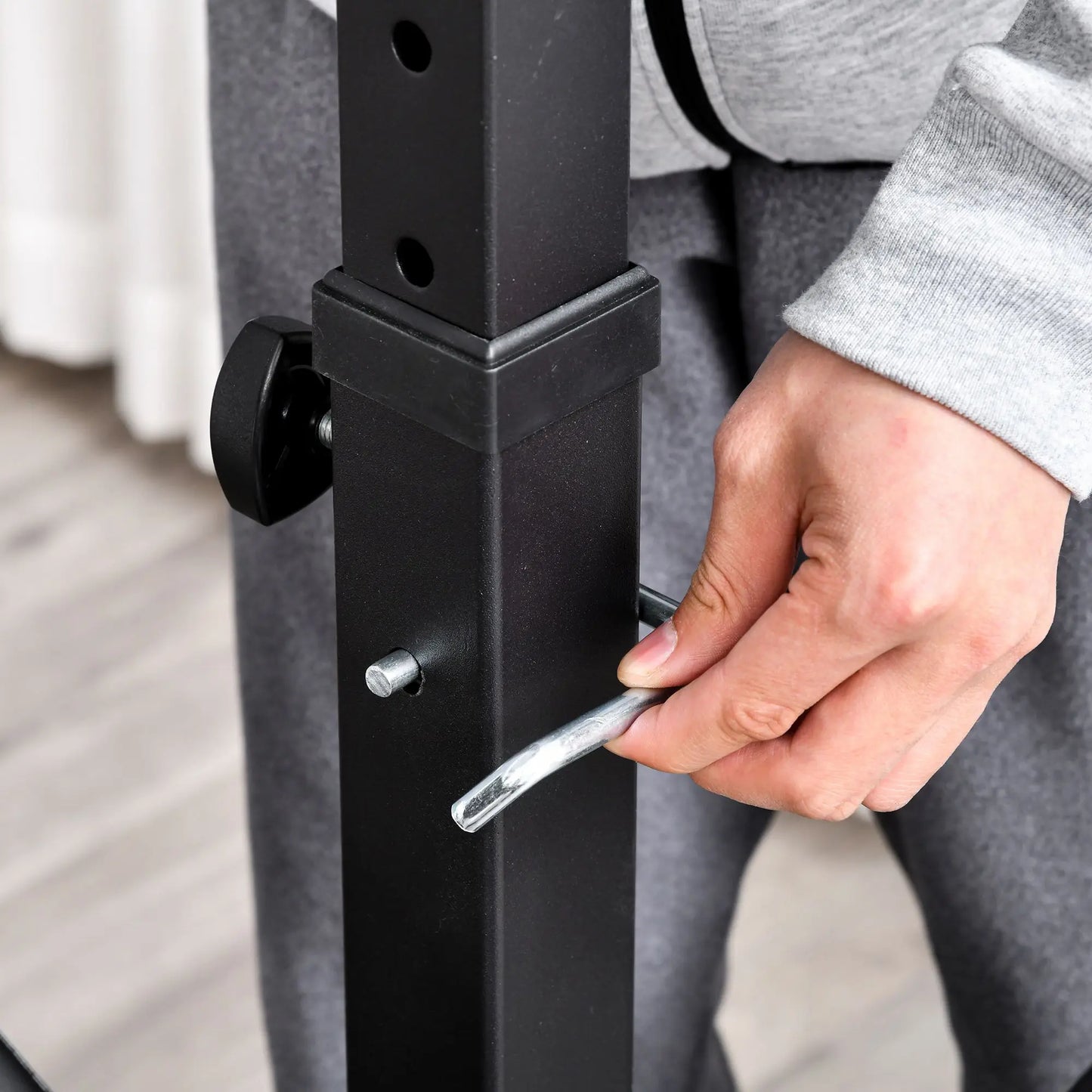 Wearing a gray sweatshirt and sweatpants, a person adjusts a metal pin on the black vertical frame of the JB Muscle HOMCOM Multi-Exercise Full-Body Weight Rack with bench press, leg extension, chest fly resistance band, and preacher curl.